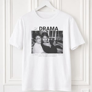 May include: A white short-sleeve t-shirt featuring a black and white photograph of two people. Above the photo is the text "The DRAMA" and below the photo is the text "I'LL SHOOT YOU FIRST. THEN YOUR PARENTS. THEN I'LL SHOOT YOU AND YOUR PARENTS."