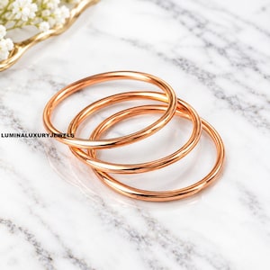 Puede incluir: Tres brazaletes de metal en tono oro rosa pulido apilados juntos. Las pulseras circulares presentan una superficie lisa y reflectante con un diseño de banda delgada y simple. El texto LUMINALUXURYJEWELS está impreso en negro a la izquierda.