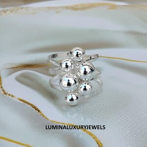 May include: A silver-toned ring featuring multiple thin, overlapping bands adorned with a cluster of smooth, polished spherical beads of varying sizes. The text LUMINALUXURYJEWELS is printed in black at the bottom of the image frame.