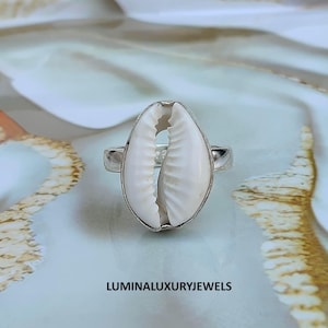 Può includere: Un anello in argento con una conchiglia di ciprea bianca al centro. La conchiglia ha una forma ovale liscia con un'apertura longitudinale centrale e bordi scanalati. La fascia metallica è in argento lucido. Il testo LUMINALUXURYJEWELS è visibile.