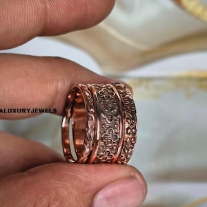 Puede incluir: Un anillo de metal en tono cobre con una banda ancha que presenta un patrón floral en relieve repetitivo y una superficie interior lisa. El texto LUMINALUXURYJEWELS es visible en el lado izquierdo del anillo, que tiene un acabado pulido.
