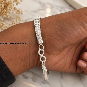 Könnte beinhalten: Ein silbernes Armband mit geflochtenem Kettenmuster und einem markanten Verschluss in Form eines Unendlichkeitssymbols. Das Metall hat eine helle, polierte Oberfläche. Der Text LUMINALUXURYJEWELS ist links im Bild neben der Kette zu sehen.