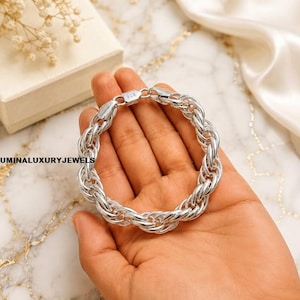 Puede incluir: Una pulsera de cadena de cuerda de plata con eslabones gruesos y entrelazados con un acabado pulido. El cierre está claramente marcado con el número 925, lo que indica una composición de plata esterlina. El texto LUMINALUXURYJEWELS es visible.