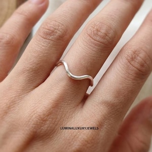 May include: A polished silver ring featuring a smooth, continuous wave-like design that curves gently across the finger. The band has a reflective metallic finish and the text LUMINALUXURYJEWELS is printed in small black letters near the bottom.