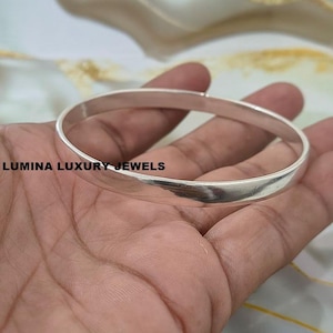May include: A polished silver bangle bracelet with a smooth, rounded surface. The text LUMINA LUXURY JEWELS is printed in black on the left side of the metal band. The bracelet features a simple, continuous circular design with a reflective finish.