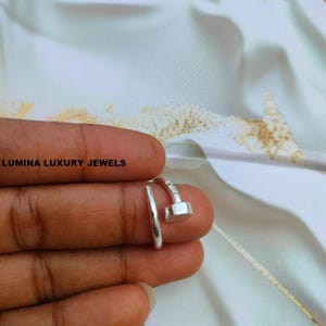 Op de afbeelding: Een zilverkleurige ring in de vorm van een spijker met een platte, ronde kop en een taps toelopend uiteinde. De metalen band is open, waardoor deze verstelbaar is. De tekst LUMINA LUXURY JEWELS is zichtbaar in de linkerbovenhoek.