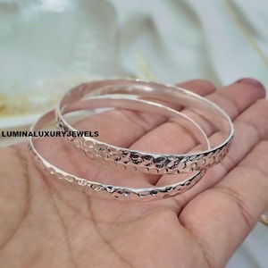 Peut inclure: Deux bracelets joncs argentés présentant une texture martelée et alvéolée sur la surface extérieure. Les bracelets ont un profil arrondi et une finition polie et réfléchissante. Le texte LUMINALUXURYJEWELS est visible sur les bracelets.