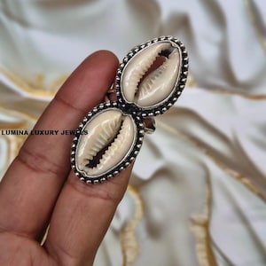 Può includere: Un anello con due conchiglie di ciprea ovali disposte verticalmente in una montatura in metallo argentato con bordo a perline. Le conchiglie sono color crema con striature naturali. Il testo LUMINA LUXURY JEWELS è visibile nell'immagine.