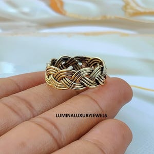 May include: A gold-toned metal ring featuring a complex, multi-strand braided design that wraps around the entire band. The surface of the metal has a polished finish with darker recessed areas. The text LUMINALUXURYJEWELS is visible below.