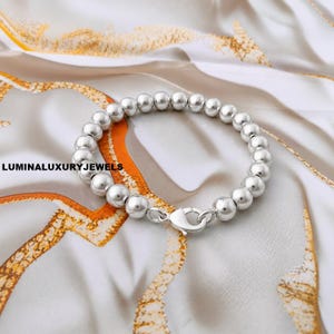 Puede incluir: Una pulsera de cuentas de plata pulida que presenta una serie de cuentas metálicas esféricas y uniformes ensartadas. La pulsera incluye un cierre de mosquetón seguro. El texto LUMINALUXURYJEWELS es visible en el lado izquierdo.