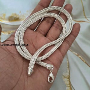 May include: A silver-toned wheat chain necklace featuring a woven, braided metal texture. The chain includes a polished metal lobster claw clasp at one end and a smooth, tapered end cap at the other. The text LUMINALUXURYJEWELS is visible on the left.