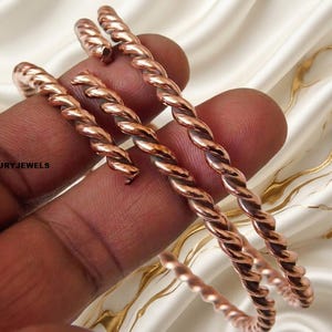 May include: Two copper-colored metal bracelets featuring a twisted rope design. The bracelets have an open-ended cuff style with rounded tips. The text LUMINALUXURYJEWELS is printed in black on the left side of the image near the metal surface.