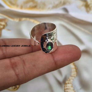 May include: A wide, open-band silver ring featuring a hammered texture and a single oval-shaped iridescent gemstone. The text LUMINA LUXURY JEWELS is printed on the left side of the image. The ring has a polished finish and a modern, bold design.