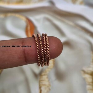 Puede incluir: Un conjunto de tres anillos de metal trenzado de color cobre. Los anillos presentan una textura continua similar a una cuerda y un acabado metálico. El texto LUMINA LUXURY JEWELS está impreso en negro en el lado izquierdo de la imagen.
