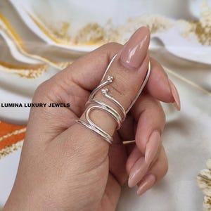 May include: A silver-toned metal ring featuring a multi-loop spiral design that wraps around the finger. The ring has two distinct ends, each finished with a small, three-bead cluster detail. The text LUMINA LUXURY JEWELS is visible on the left.