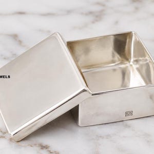 Puede incluir: Una caja rectangular de plata pulida con la tapa separada, que presenta una superficie lisa y reflectante. El texto 925 está estampado en el lateral de la caja y el texto LUMINALUXURYJEWELS es visible en el lado izquierdo de la tapa.