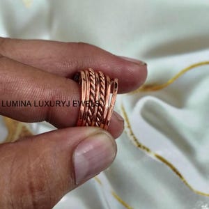 Op de afbeelding: Een koperkleurige ring bestaande uit vijf individuele banden, waaronder drie gladde banden en twee gedraaide touwachtige banden. De tekst LUMINA LUXURYJ EWELS is zichtbaar in het midden van de ring. Het metaal heeft een warme afwerking.