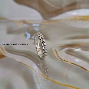 Puede incluir: Un anillo de tono plateado que presenta un patrón continuo en relieve de trigo u hojas alrededor de toda la banda. La superficie metálica tiene un acabado pulido con áreas hundidas oscurecidas para resaltar la textura. El texto LUMINALUXURYJEWELS es visible.