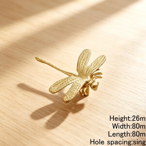 Peut inclure: Un bouton de meuble en m&eacute;tal dor&eacute; en forme de libellule avec des textures d'ailes d&eacute;taill&eacute;es. Le bouton dispose d'un point de fixation &agrave; trou unique. Le texte indique une hauteur de 26mm, une largeur de 80mm, une longueur de 80mm.