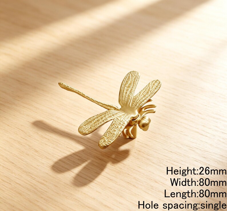 Peut inclure: Un bouton de meuble en m&eacute;tal dor&eacute; en forme de libellule avec des textures d'ailes d&eacute;taill&eacute;es. Le bouton dispose d'un point de fixation &agrave; trou unique. Le texte indique une hauteur de 26mm, une largeur de 80mm, une longueur de 80mm.