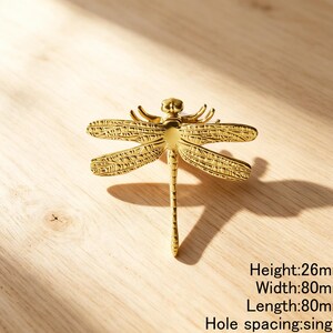 Peut inclure: Un bouton de meuble dor&eacute; en forme de libellule avec des ailes textur&eacute;es et un corps lisse. La quincaillerie pr&eacute;sente un espacement de trou unique et des dimensions de 26 mm de hauteur, 80 mm de largeur et 80 mm de longueur indiqu&eacute;es par le texte.