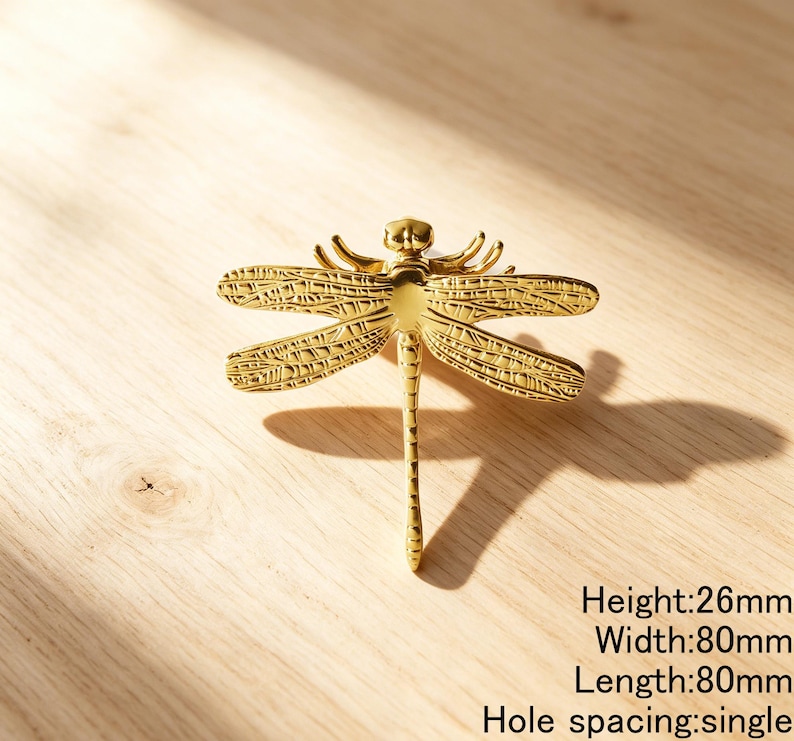 Peut inclure: Un bouton de meuble dor&eacute; en forme de libellule avec des ailes textur&eacute;es et un corps lisse. La quincaillerie pr&eacute;sente un espacement de trou unique et des dimensions de 26 mm de hauteur, 80 mm de largeur et 80 mm de longueur indiqu&eacute;es par le texte.