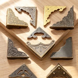 Pode incluir: Uma coleção de nove cantoneiras de metal com vários acabamentos em bronze, latão e metal escuro. Cada peça apresenta padrões intrincados em relevo, incluindo motivos florais e volutas, com pequenos orifícios pré-perfurados.