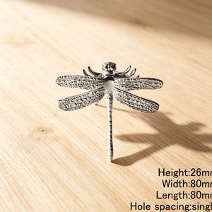 Peut inclure: Un bouton de meuble en forme de libellule de couleur argent&eacute;e avec des d&eacute;tails d'ailes complexes. La quincaillerie pr&eacute;sente une hauteur de 26 mm, une largeur de 80 mm, une longueur de 80 mm et un espacement de trou unique pour le montage.