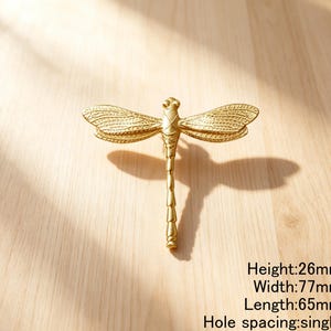 Peut inclure: Un bouton de meuble dor&eacute; en forme de libellule avec des ailes textur&eacute;es et un corps segment&eacute;. La quincaillerie pr&eacute;sente une hauteur de 26mm, une largeur de 77mm, une longueur de 65mm et un espacement de trou unique pour le montage.