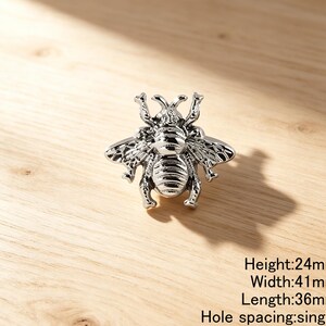Peut inclure: Un bouton de meuble en forme d'abeille argent&eacute;e avec des ailes et des segments de corps d&eacute;taill&eacute;s. La quincaillerie pr&eacute;sente une conception &agrave; trou de montage unique. Le texte indique une hauteur de 24mm, une largeur de 41mm, une longueur de 36mm.