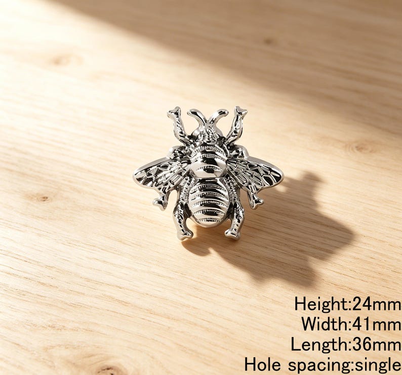 Peut inclure: Un bouton de meuble en forme d'abeille argent&eacute;e avec des ailes et des segments de corps d&eacute;taill&eacute;s. La quincaillerie pr&eacute;sente une conception &agrave; trou de montage unique. Le texte indique une hauteur de 24mm, une largeur de 41mm, une longueur de 36mm.
