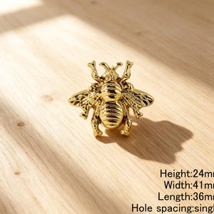 Peut inclure: Un bouton de meuble dor&eacute; en forme d'abeille avec des ailes textur&eacute;es et un abdomen ray&eacute;. La quincaillerie pr&eacute;sente une fixation &agrave; trou unique. Le texte indique une hauteur de 24mm, une largeur de 41mm, une longueur de 36mm et un espacement unique.