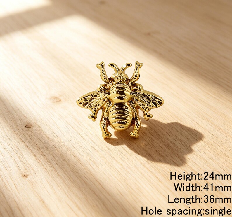 Peut inclure: Un bouton de meuble dor&eacute; en forme d'abeille avec des ailes textur&eacute;es et un abdomen ray&eacute;. La quincaillerie pr&eacute;sente une fixation &agrave; trou unique. Le texte indique une hauteur de 24mm, une largeur de 41mm, une longueur de 36mm et un espacement unique.