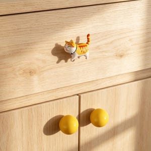 Puede incluir: Un tirador de cajón con forma de pequeño gato atigrado naranja y blanco con collar negro, situado sobre dos pomos de armario redondos y amarillos en una superficie de madera clara con un cajón horizontal y dos puertas de armario inferiores.