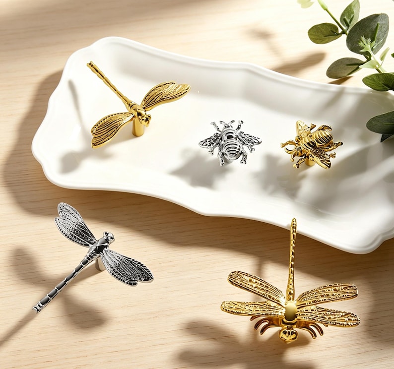 Peut inclure: Un ensemble de cinq boutons de placard en m&eacute;tal en forme d'insectes, comprenant trois libellules et deux abeilles. Le lot pr&eacute;sente un m&eacute;lange de finitions dor&eacute;es et argent&eacute;es polies, avec des ailes et des corps d&eacute;taill&eacute;s, pos&eacute;s sur un plateau blanc.