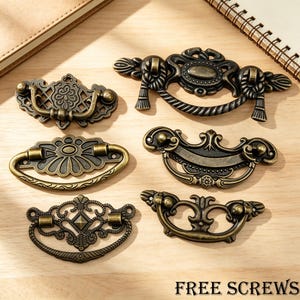 May include: A collection of six antique brass-toned metal drawer pulls featuring ornate, intricate filigree and scrollwork designs with swinging handles. The hardware displays various decorative shapes and patterns, and the text FREE SCREWS is visible.
