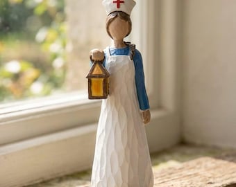 May include: A figurine of a nurse wearing a blue dress and a white apron with a textured finish. The figure wears a white cap with a red cross symbol and holds a small, yellow-lit lantern in one hand. The sculpture has a carved appearance.