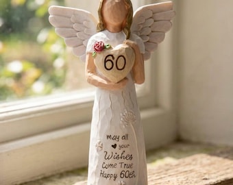 Può includere: Una statuetta di angelo in piedi con capelli castano chiaro e ali bianche, che indossa un lungo abito bianco. L'angelo tiene un oggetto a forma di cuore con il numero 60. L'abito riporta il testo: May all your Wishes Come True Happy 60th.