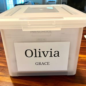 May include: A clear plastic storage bin with a matching lid, featuring a white rectangular label on the front. The label displays the name Olivia in a large serif font, with the name GRACE printed in a smaller sans-serif font below it.