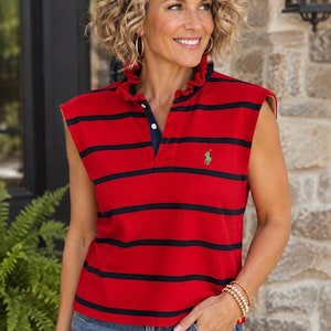 May include: A sleeveless red polo shirt featuring horizontal black stripes, a ruffled collar, and a partial button placket. A small green embroidered logo is visible on the left chest area. The shirt is paired with blue denim jeans.