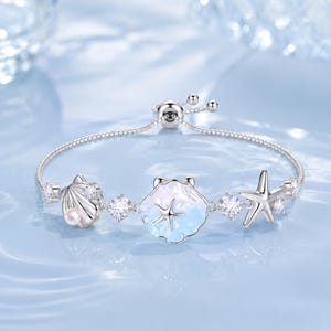 May include: A silver-toned adjustable bracelet featuring a central shell charm with a blue and white gradient finish, accented by a small starfish. The chain includes a pearl-adorned shell, a starfish charm, and several clear, sparkling round stones.