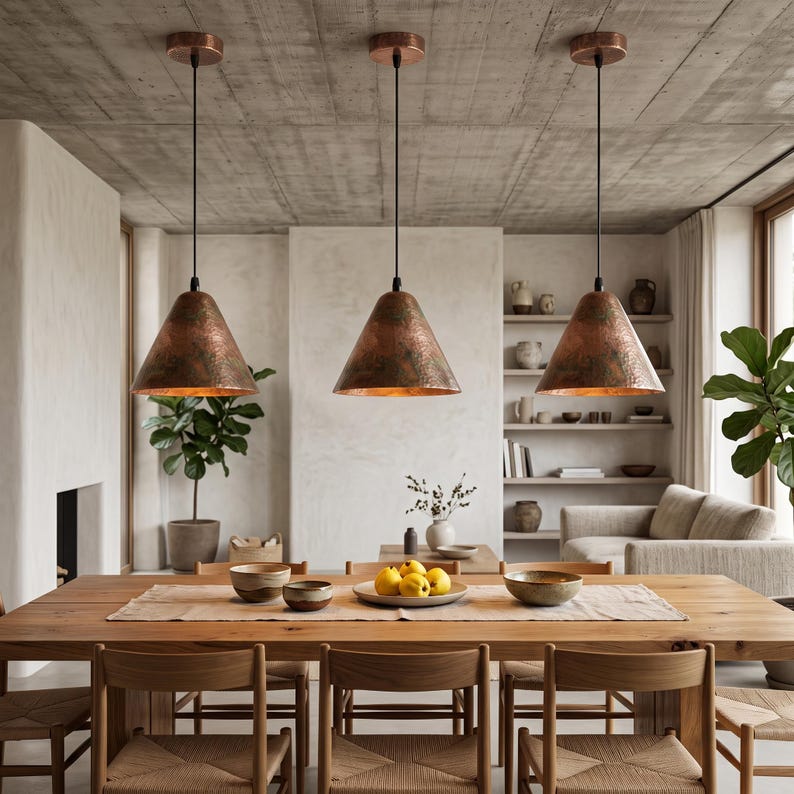 May include: Three cone-shaped pendant lights with a hammered copper finish hang from black cords above a long wooden dining table. The fixtures feature a textured metallic surface and are suspended from circular copper ceiling mounts.