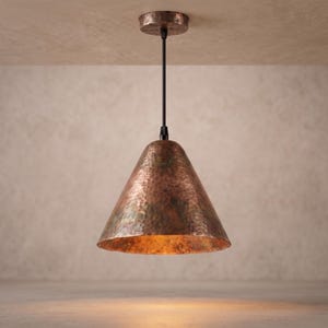 May include: A cone-shaped pendant light fixture featuring a hammered copper finish with a warm, metallic texture. The lamp is suspended by a thin black cord from a matching circular copper ceiling canopy, casting a soft, downward glow.