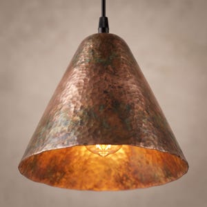 May include: A conical pendant light fixture featuring a hammered copper exterior with a weathered, oxidized green patina finish. The shade is suspended by a black cord and houses a single glowing filament bulb visible within the wide opening.