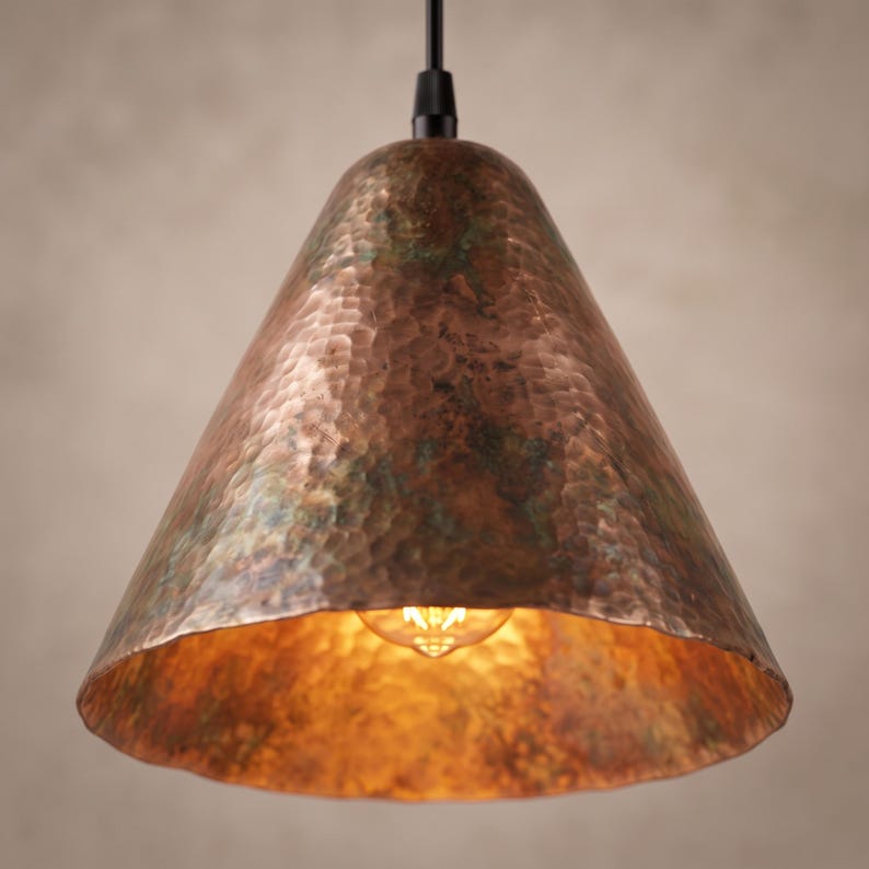 May include: A conical pendant light fixture featuring a hammered copper exterior with a weathered, oxidized green patina finish. The shade is suspended by a black cord and houses a single glowing filament bulb visible within the wide opening.
