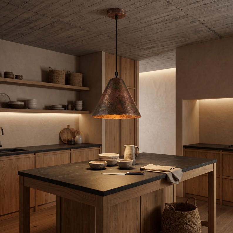 May include: A conical pendant light fixture with a textured, dark copper-toned metal exterior and a warm, reflective interior finish. The lamp is suspended by a thin black cord from a matching circular ceiling mount, casting a soft downward glow.