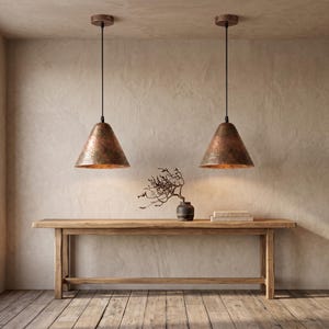May include: Two cone-shaped pendant lights with a hammered copper finish suspended by black cords from the ceiling. The lights hang above a long, rectangular wooden console table featuring a dark vase with bare branches and a stack of books.