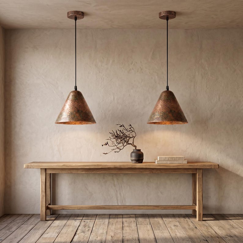 May include: Two cone-shaped pendant lights with a hammered copper finish suspended by black cords from the ceiling. The lights hang above a long, rectangular wooden console table featuring a dark vase with bare branches and a stack of books.