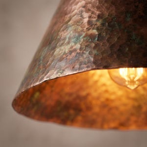May include: A dome-shaped pendant light fixture featuring a hammered copper exterior with a weathered, oxidized green patina finish. The interior surface reflects the warm, golden glow of an exposed filament light bulb housed within the shade.