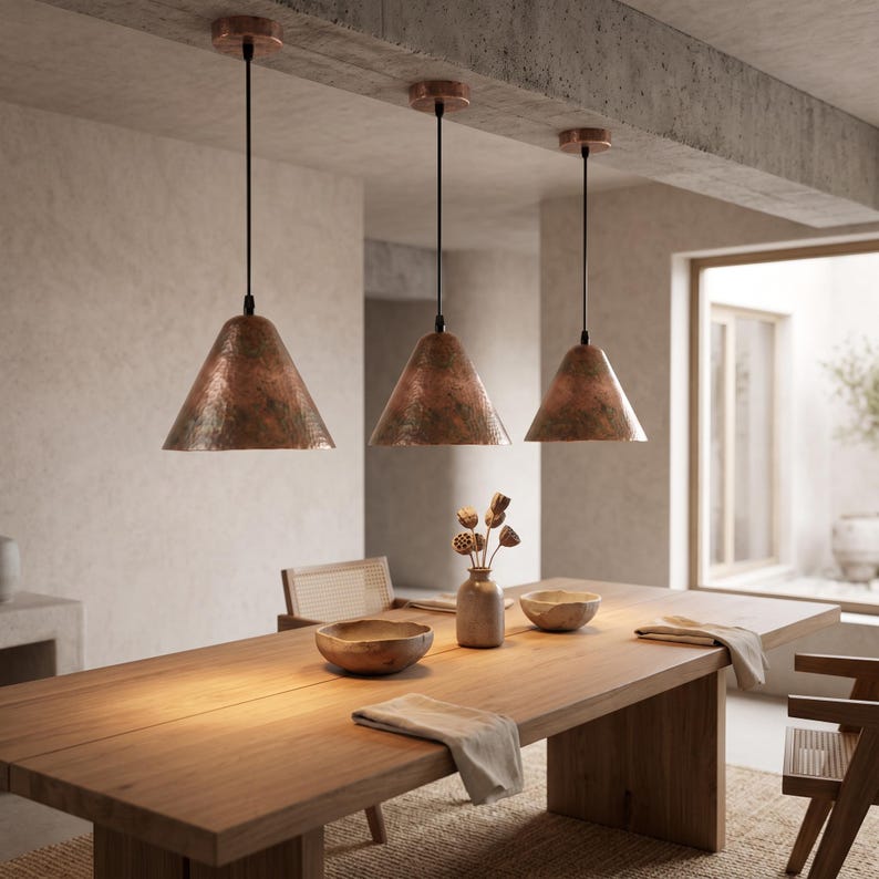 May include: Three cone-shaped pendant lights with a hammered copper finish suspended by black cords from a concrete ceiling. Each light fixture features a metallic, textured surface that reflects light, hanging in a row above a wooden table.